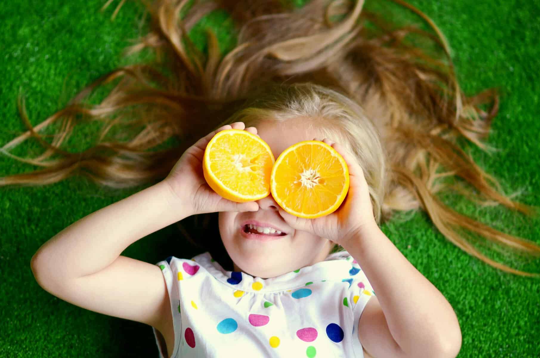 A cheerful girl lying on grass with orange slices, depicting playfulness and summer joy.